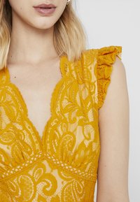 Gul spetsblus med volangärmar, med intrikata blommönster och djup V-ringning. Lätt, genomskinlig tygdesign.