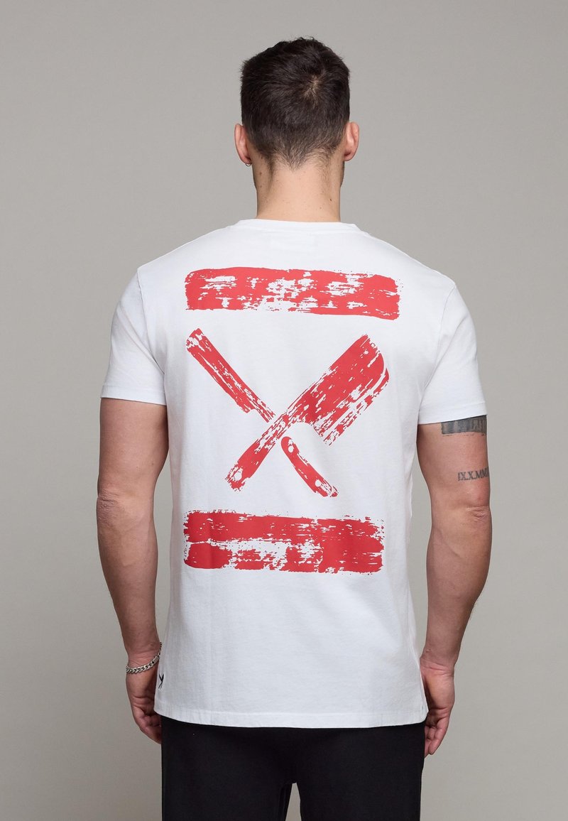 Homme aux cheveux courts et foncés, avec un tatouage sur le bras, portant un T-shirt blanc orné au dos d'illustrations rouges usées de hachoirs croisés.