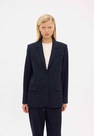 Femme blonde portant un blazer bleu marine à deux boutons et un pantalon assorti, debout devant un fond clair uni.