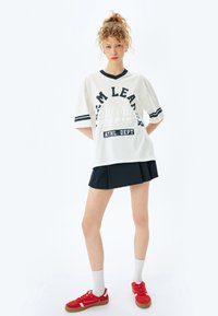 Hvid oversized t-shirt med marineblå accenter og tekst, parret med en sort plisseret nederdel. Modellen har røde sneakers og hvide ankelsokker på.