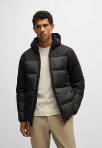 Man wearing a black padded jacket with hood, beige sweater, and light brown pants, standing against a plain light background.