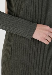 Vêtement à manches longues côtelé en tissu vert olive foncé. Présente des rayures verticales et une finition texturée. Main posée sur le tissu.