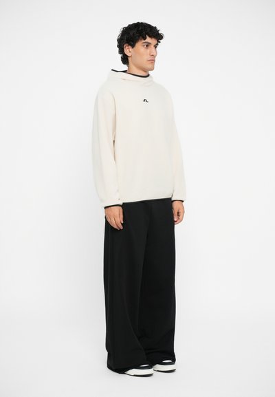 Hellbeige, übergroßer Pullover mit hohem Kragen und Logo auf der Brust. Kombiniert mit lässigen, schwarzen Hosen und schwarz-weißen Sneakers.