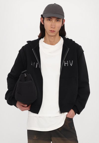 Black zip-up hoodie with white star pattern, worn over a white T-shirt, paired with black bag and gray cap, standing against a white background.