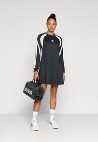 Juoda sportinė suknelė ilgomis rankovėmis, baltomis detalėmis ir Adidas logotipu. Derinama su baltomis kojinėmis ir sportbačiais, rankose laikomas juodas sportinis krepšys.