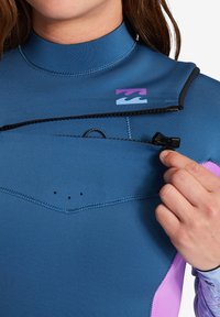 Pianka o niebieskim materiale neoprenowym z różowymi akcentami, z zamkiem błyskawicznym z przodu i wysokim kołnierzem. Wyraźne logo w purpurowym i niebieskim kolorze nad zamkiem.