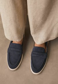 Navy blue suede loafers with white rubber soles, featuring a stitched detailing and wide strap across the top, set against beige fabric.