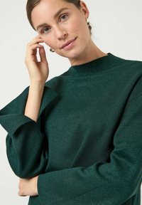 Ciemnozielony sweter o fakturowanej powierzchni z wysokim kołnierzem i szerokimi, złożonymi rękawami. Materiał ma subtelny połysk. Akcenty obejmują złote kolczyki.