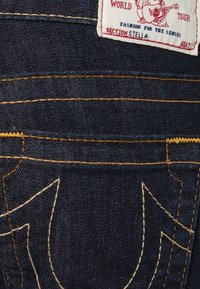 Denimjeans i mörkblå färg med gula sömmar. Inkluderar en vit etikett med röd tryck och en broderad design på bakfickan.