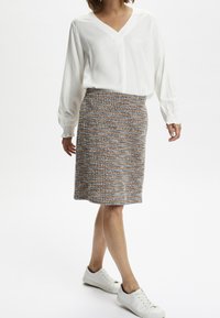 Blouse blanche à col en V avec des manches froncées, associée à une jupe en tissu tissé multicolore mi-longue. Le mannequin porte des baskets blanches.