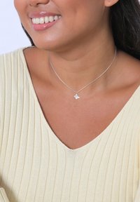 Sølv halskæde med en lille abstrakt vedhæng. Båret over en ribbet, lys gul v-neck top. Glat tekstur, minimalistisk design.