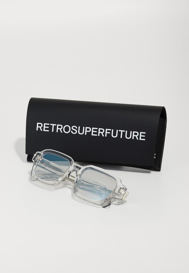 Lunettes rectangulaires transparentes avec des verres bleu clair, reposant sur un étui noir portant le texte blanc "RETROSUPERFUTURE." Matériau lisse et design moderne.