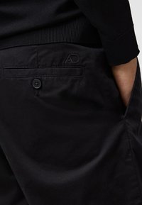 Pantaloni neri con una texture liscia, dotati di una tasca posteriore, chiusura con bottone e logo ricamato discreto. Vita a maglia con dettaglio a coste.