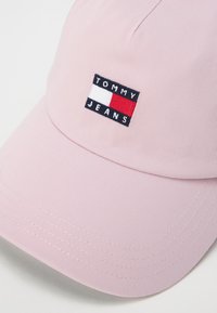 Pink cap with textured fabric, featuring a rectangular embroidered logo in black, white, and red that says "TOMMY JEANS." Curved brim.