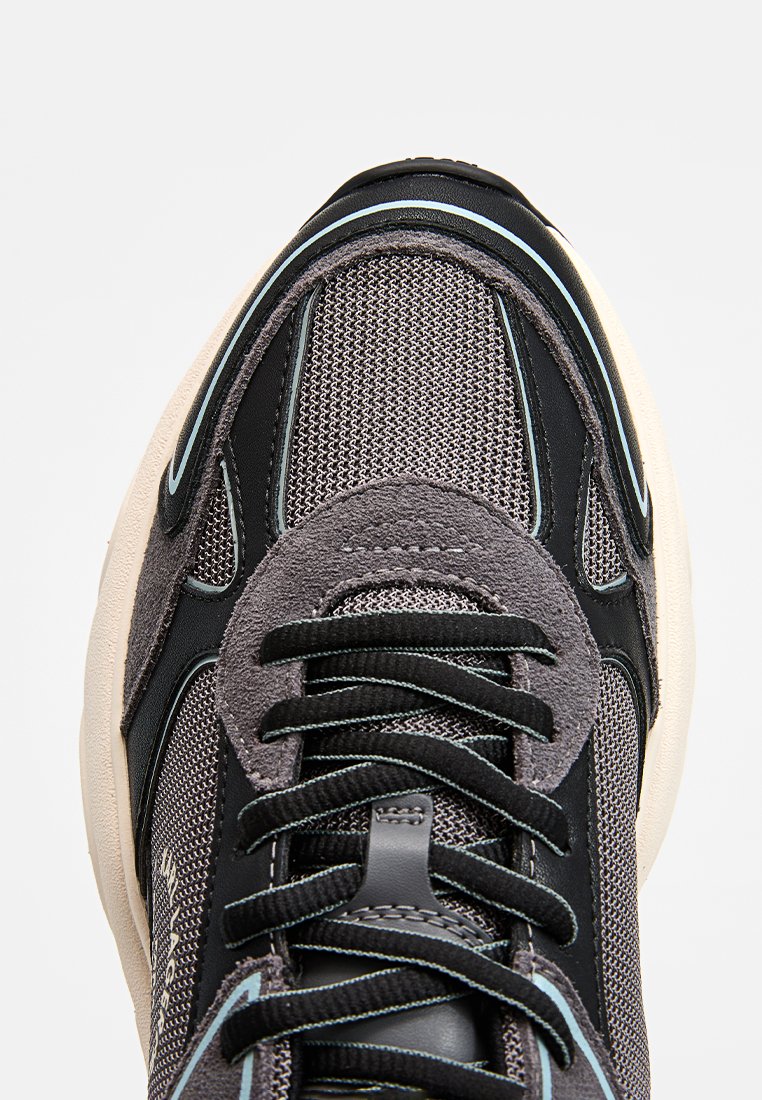 Zapatilla gris con parte superior de malla y ante, acentos negros y cordones texturizados. Punta redondeada y suela acolchada con un detalle de costura distintivo.