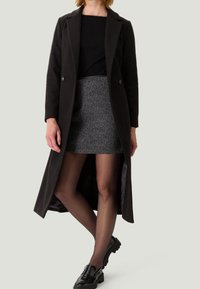 Manteau long noir à la coupe ajustée, associé à une mini-jupe grise à motifs. Le mannequin porte des collants noirs transparents et des mocassins noirs épais.