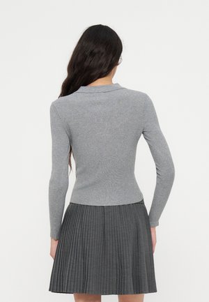 Femme aux longs cheveux foncés portant un pull côtelé gris cintré et une jupe plissée gris foncé, vue de dos sur un fond uni.