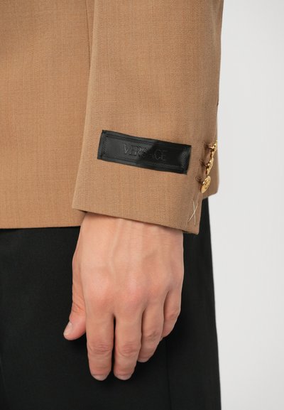 Manga de saco marrón con una etiqueta negra de Versace, botones dorados y detalles de costura sutil. Textura de tela suave y diseño entallado.