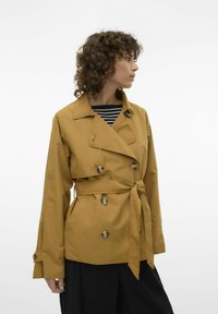 Sennepsgul trenchcoat med dobbeltknapning, store knapper, en bælte i taljen og lange ærmer, båret over en stribet top.