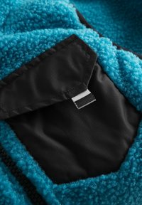 Blaues Fleece-Gewebe mit strukturierter Oberfläche, ausgestattet mit einer schwarzen Tasche und einem kontrastierenden schwarz-weißen Akzentstreifen.