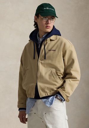 Beige bomberjakke med navyblåt hætteforet, lynlåslukning, sidelommer og broderet logo. Grøn baseballkasket med tekstdetalje.