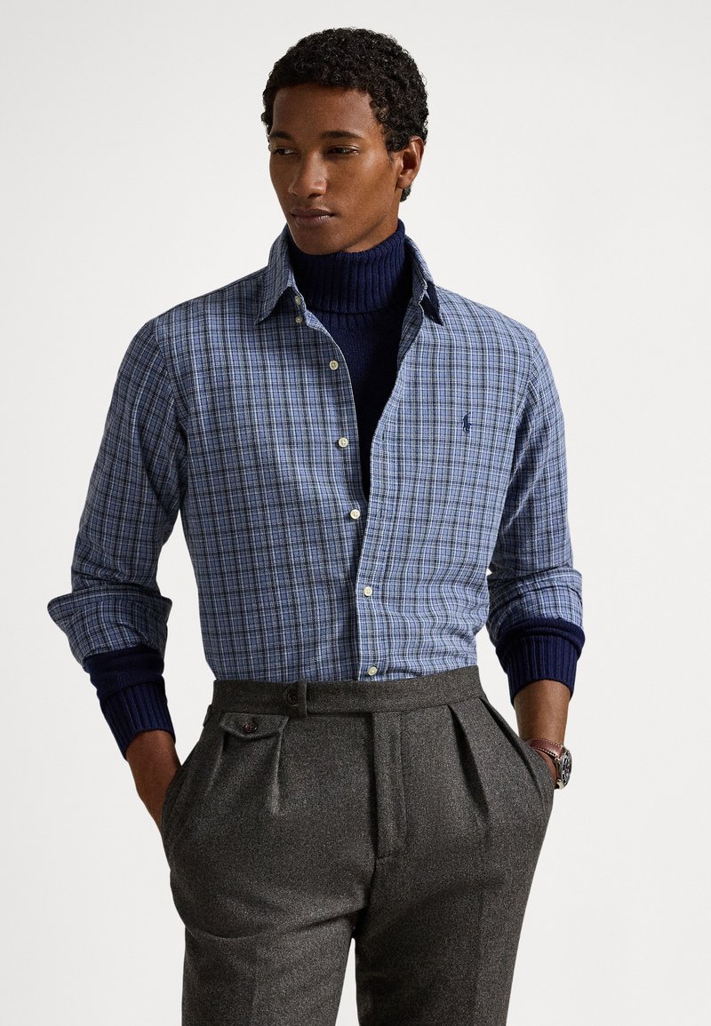 Mėlyna languota marškiniai su sagomis ant apykaklės, derinami su tamsiai mėlynu golfo megztiniu ir tamsiai pilkomis klostuotomis kelnėmis. Matomi riešo rankogaliai ir laikrodis.