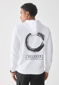 Uomo che indossa un hoodie bianco con un design a pennellata circolare nera e il testo "BALANCE" sulla schiena, in piedi di fronte a uno sfondo bianco semplice.