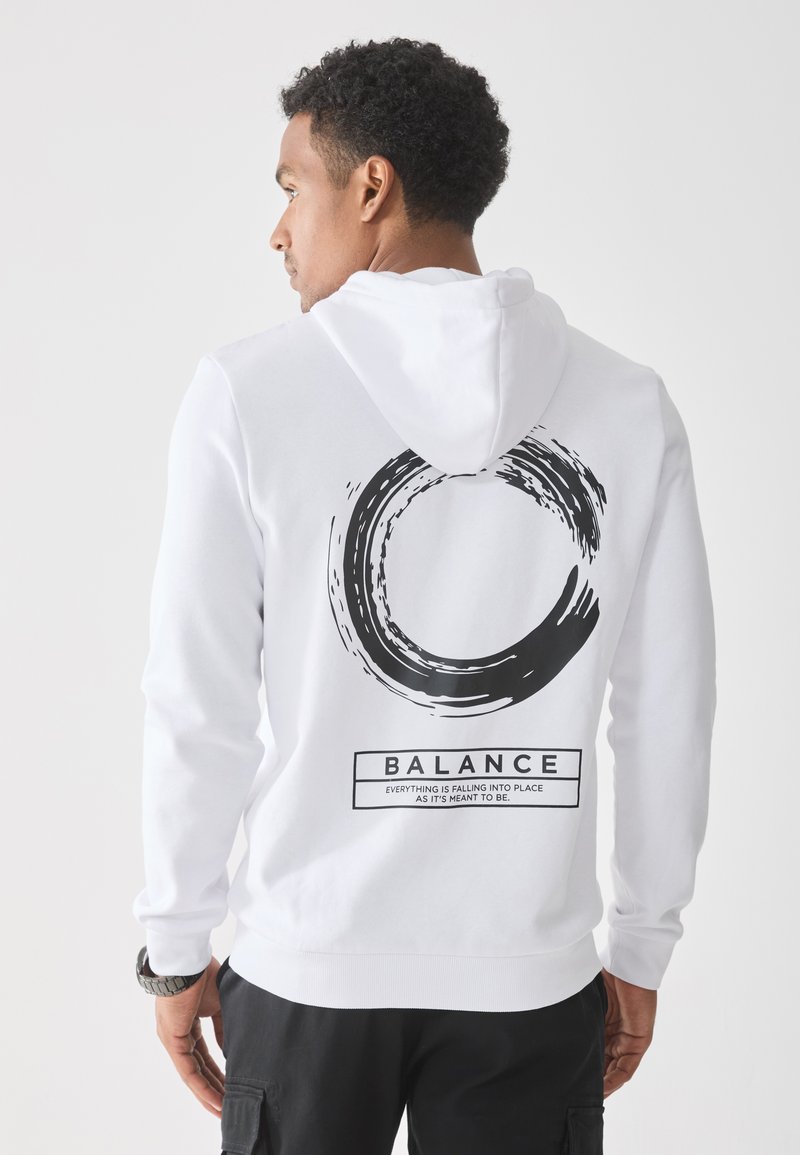 Uomo che indossa un hoodie bianco con un design a pennellata circolare nera e il testo "BALANCE" sulla schiena, in piedi di fronte a uno sfondo bianco semplice.