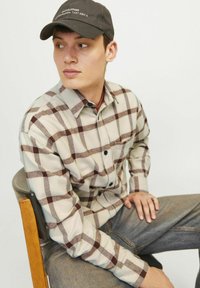 Chemise à carreaux crème et marron, avec un col boutonné et une poche sur la poitrine, associée à un jean en denim foncé et une casquette noire.