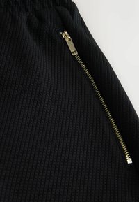 Tissu noir texturé avec une fermeture éclair dorée partiellement ouverte, montrant un motif côtelé vertical et une ouverture de poche.