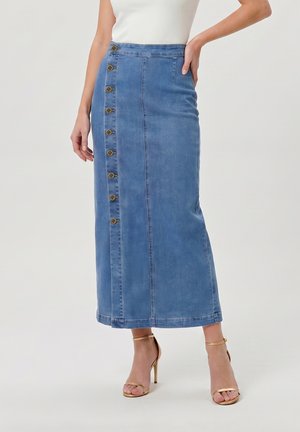 Femme portant une jupe midi en denim bleu taille haute avec des boutons sur le côté et des sandales à talons hauts à bride cheville dorées.