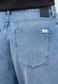 Ljusblå denimjeans har en slät textur med en lädermärkesskylt och en liten etikett på bakfickan. Klassisk femficksdesign.