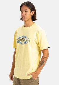 Camiseta de algodón amarilla con un estampado gráfico que presenta hojas de palmera azules y blancas y texto en negro. Corte relajado y mangas cortas.