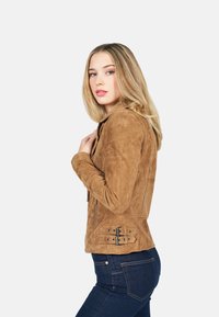 Bruine suède jas met een getailleerd ontwerp, ritssluitingen, zijzakken en dubbele gespenaccenten op de taille. Gedragen over donkere denim.