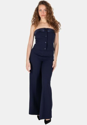 Donna con capelli ricci che indossa un top blu navy senza spalline con bottoni e pantaloni a gamba larga coordinati, in piedi con le braccia rilassate lungo i lati.