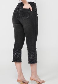 Crne traperice capri dužine s vezom cvjetnog uzorka na rubu, izrezanim rubovima i uskim krojem. Ima dvije stražnje džepove.