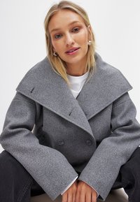 Grå ullblandad dubbelknäppt kappa med breda slag, stora knappar och överdimensionerade ärmar. Slät textur med subtila sömdetaljer.