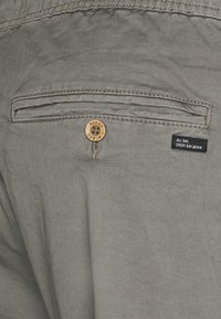 Pantaloni bej cu un singur buzunar pe spate, cu închidere cu nasture din lemn și o etichetă din material textil negru cu inscripția "ALL DAY EVERY DAY DENIM". Textură netedă.
