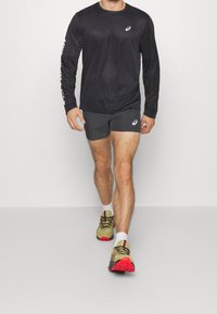 Langärmliges schwarzes Sportshirt mit Logo, graue Shorts und mehrfarbige Laufschuhe mit roten Sohlen. Person läuft vorwärts.