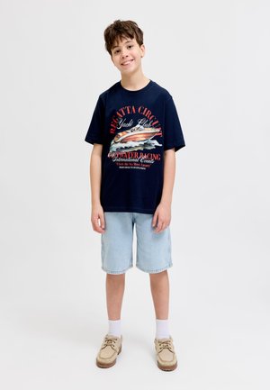 Naeratav poiss, kes kannab tumesinist t-särki laevamustriga, heledaid teksapükse, valgeid sokke ja beeži vabaajajalatsit, seisab valgel taustal.