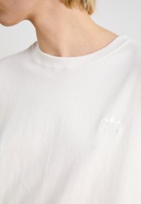 Bijela pamučna majica s rebrastim okovratnikom i izvezenim adidas logom u suptilnom toniranom dizajnu na lijevoj strani prsnog dijela.