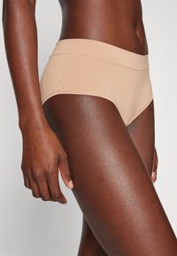 Beige seamless hipster underwear with a wide waistband, made from a smooth, stretchy fabric, displayed on a model against a neutral background.