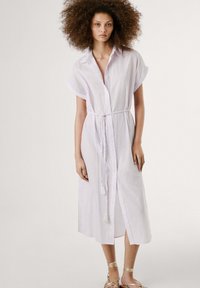 Vit randig skjortklänning med korta uppvikta ärmar, midjeband och sidokrossningar, tillverkad av lättviktigt tyg. Silverfärgade sandals med remmar.