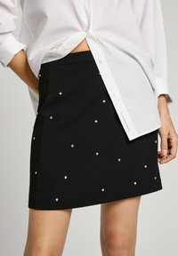 Gonna nera con decorazioni in argento sparse, silhouette sartoriale e una texture liscia, abbinata a una camicia bianca con bottoni.