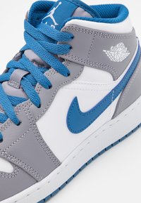 Baskets montantes en gris, blanc et bleu. Caractéristiques en cuir lisse, lacets coordonnés en bleu, et un logo aile proéminent sur le côté.