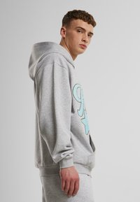 Pelēka kapuci svītrainā krekla ar lieliem tirkīza izšūtiem burtiem un zvaigžņu dizainu. Mīksta tekstūra un ribošanas aproces un apakšdaļas.