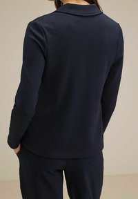 Person i en marinblå långärmad skjorta med krage och matchande marinblå byxor, som står med händerna i fickorna, med ryggen vänd mot betraktaren.