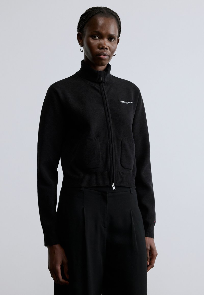 Fekete cipzáras pulóver magas gallérral, bordázott szegéllyel és elülső zsebekkel. Diszkrét márkajelzés a mellkason, puha tapintású anyag és testhez simuló szabás jellemzi.