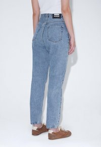 Světlé modré džíny s vysokým pasem a rovnými nohavicemi, vybavené dvěma zadními kapsami a černou značkovou nášivkou na pase.