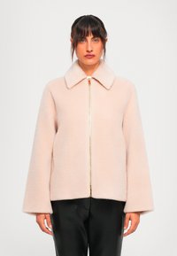Tume juustega naine kannab heledat roosa karvast zip-up jope, millel on laiad varrukad ja musti pükse, seistes ühtlasel valgel taustal.